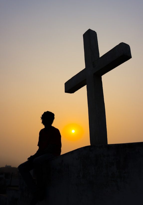 silhouette of man sitting on the edge of a cross during sunset
